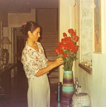 Una mujer con moño en el cabello y camisa floral arregla un ramo de rosas rojas en un jarrón a rayas. De fondo se ven escaleras y una lámpara