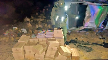 Vista nocturna de un oficial con linterna inspeccionando cajas y una pila de libros de colores, con un vehículo blanco volcado parcialmente al fondo entre vegetación
