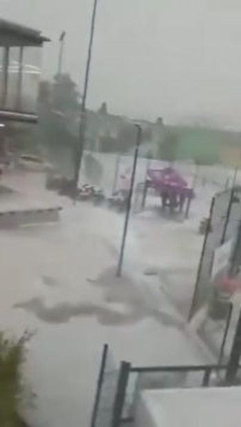 Una calle anegada con agua cubriendo el suelo, edificios de dos pisos, árboles, mesas y sillas al aire libre, y postes