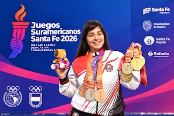Una mujer sonriente viste ropa deportiva con medallas al cuello y sostiene un peluche y más medallas frente a un panel publicitario azul