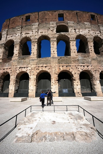 Vista del Coliseo en Roma con su fachada de piedra y arcos contra un cielo azul brillante. En primer plano, tres personas caminan sobre un suelo de concreto y grava