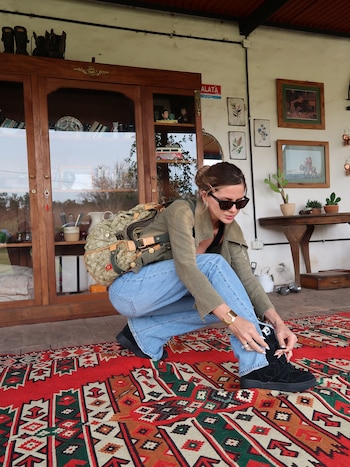 Una mujer con gafas de sol, chaqueta verde y pantalones vaqueros se agacha sobre una alfombra roja para ajustarse los zapatos