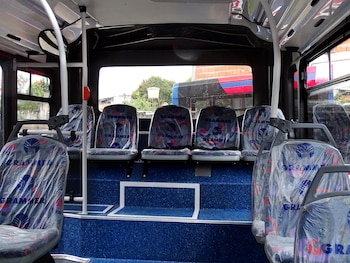 Interior de un minibús con asientos nuevos cubiertos en plástico transparente y suelo azul. Por las ventanas se aprecia parte de otro vehículo y edificios