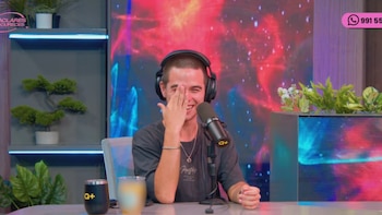 Un hombre con auriculares se tapa el rostro con una mano frente a un micrófono en un plató con pantallas e iluminación de colores