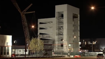 Vista nocturna de un edificio de construcción moderna con múltiples pisos y aberturas, situado junto a una grúa de torre y bajo luces de calle brillantes