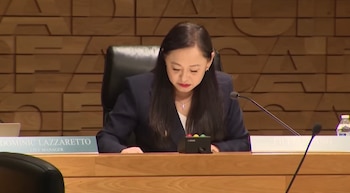 Eileen Wang, una mujer de cabello oscuro, viste blazer azul marino y blusa blanca, mirando hacia abajo en una mesa de madera con un micrófono y un dispositivo