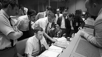 Un grupo de diez hombres vestidos con camisas y corbatas se agrupan alrededor de un escritorio en un cuarto de control, revisando papeles y equipos