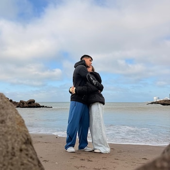 Pareja joven abrazándose en una playa arenosa junto al mar, con el cielo nublado. Rocas en la izquierda y edificios lejanos a la derecha