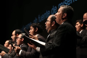 Primer plano de siete hombres en un coro con trajes oscuros, camisas blancas, y carpetas negras. Tienen la boca abierta al cantar, con un fondo oscuro