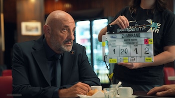 Primer plano de Gabriel Goity, un hombre calvo con barba y traje oscuro, sentado en una mesa de restaurante con una claqueta de Disney+ sostenida a su lado