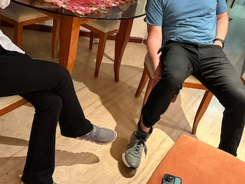 Dos personas sentadas junto a una mesa de madera y vidrio; una de ellas lleva un dispositivo electrónico negro en el tobillo derecho