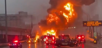 Vista aérea de una explosión masiva con llamas intensas y humo denso sobre una autopista, rodeada de varios camiones de bomberos y vehículos policiales con luces intermitentes