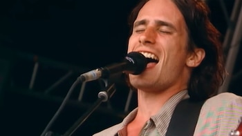 Primer plano de Jeff Buckley cantando en un micrófono con los ojos cerrados, la boca abierta, pelo castaño largo y una camisa a rayas