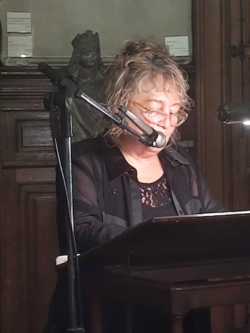 Angela Pradelli, mujer de pelo rubio rizado y gafas, habla en un micrófono en un atril de madera, con una estatua oscura y paneles de madera de fondo