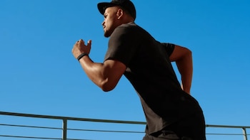 Hombre musculoso de perfil corre hacia la derecha con camiseta y gorra negras, y banda de actividad, bajo un cielo azul claro con una barandilla metálica abajo