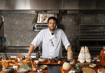 Hombre vestido de chef con gafas en el pecho, de pie en una cocina profesional con encimera de piedra. Sobre la mesa, mariscos, hierbas y piezas de cerámica