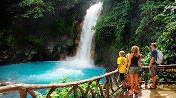 Cuatro personas observan una cascada blanca que cae sobre un río turquesa. Hay vegetación verde, rocas oscuras y una valla de madera.