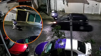 Vista aérea nocturna de una calle mojada. Una persona con paraguas dentro de un círculo naranja. Un automóvil oscuro en movimiento. Varios autos estacionados y edificios