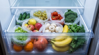 Un cajón transparente de refrigerador contiene manzanas, bananas, tomates, uvas, pepinos, brócoli y champiñones en bolsas de plástico.