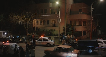 Un coche policial y una furgoneta forense con luces encendidas, personas, cinta amarilla de seguridad, un poste de luz, y un edificio de noche