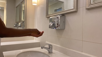 Manos extendidas bajo un secador eléctrico cromado en la pared de un baño con espejo y lavabo.