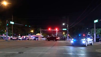 Vista nocturna de una calle con una patrulla de policía con luces intermitentes en primer plano, tráfico y señales luminosas, y tiendas al fondo