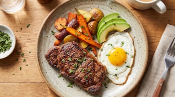 Un plato cerámico con un trozo de carne asada, un huevo frito, láminas de palta, zanahorias y papas sobre una mesa de madera.