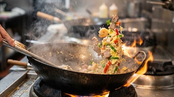 Primer plano de arroz chafua salteándose en un wok sobre llamas. Se ven gambas, carne, arroz y vegetales, con vapor ascendiendo y una mano sujetando una espátula.