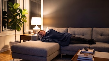 Hombre durmiendo en un sofá gris con manta azul, almohada, mesa auxiliar con lámpara y smartphone, planta en maceta, ventana, mesa de centro con libros y mando.