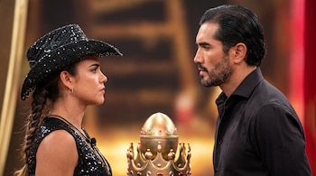 Una mujer con sombrero vaquero y un hombre con barba se miran de frente junto a una corona y un huevo dorado.