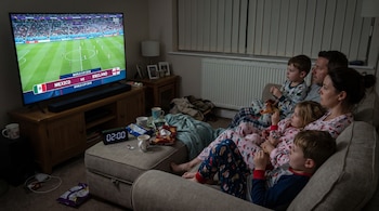 Familia de cinco personas, dos adultos y tres niños, con pijama en un sofá, ve un partido de fútbol en televisión. El reloj digital marca 02:00.