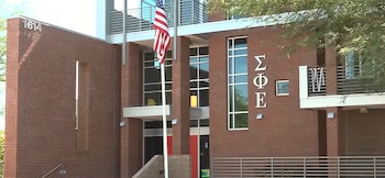 Fachada de un edificio de ladrillo con letras griegas de Sigma Phi Epsilon, una bandera de Estados Unidos y barandas metálicas