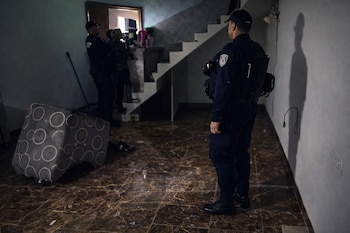 Un oficial de policía uniformado de espaldas, con chaleco táctico. Otros dos agentes y una persona al fondo. Mueble volcado. Escalera y piso con baldosas.