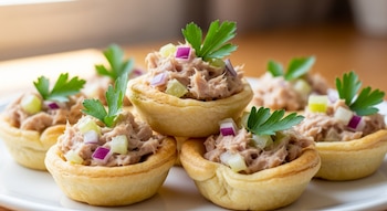 Primer plano de varias canastitas doradas rellenas de ensalada de atún con trozos de cebolla morada, apio y hojas de perejil fresco, sobre un plato blanco.