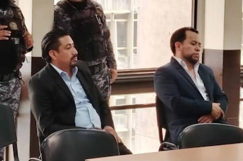 Dos hombres sentados en sillas, uno con traje azul y camisa clara, otro con traje negro y camisa azul. Detrás, dos figuras con uniforme de camuflaje