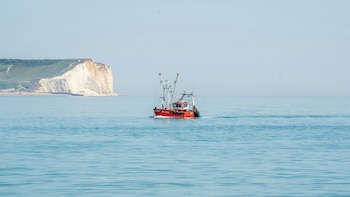 Barco pesquero rojo navegando en el mar azul con un acantilado blanco visible al fondo a la izquierda