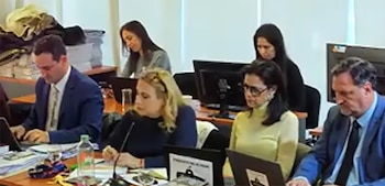Seis personas sentadas en una reunión de trabajo con mesas de madera clara. Varios usan laptops mientras otros observan, con monitores y documentos