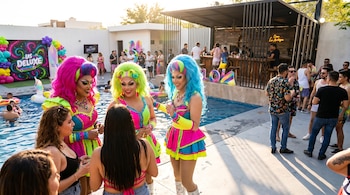 Dos drag queens con pelucas y trajes fucsia, verde neón y azul eléctrico, sonríen e interactúan con invitados en una fiesta junto a una piscina.