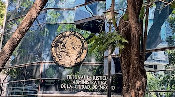 Fachada de vidrio de un edificio con el escudo nacional de México, la inscripción del Tribunal de Justicia Administrativa y árboles al frente.