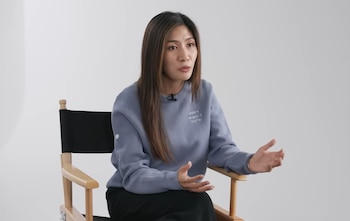 Una mujer de cabello largo con suéter azul habla sentada en una silla de director con estructura de madera y tela negra
