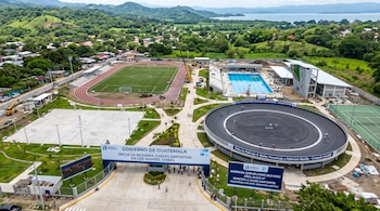 Vista aérea de un complejo deportivo con pista de patinaje circular, campo de fútbol, piscina y letreros del gobierno.