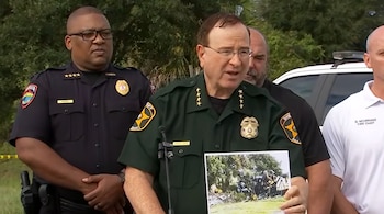 Grady Judd sostiene una fotografía impresa de un vehículo accidentado mientras habla ante los micrófonos rodeado por personal policial