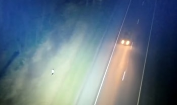 Vista aérea nocturna de una carretera oscura con un vehículo de faros brillantes en el carril derecho y una persona caminando a un lado