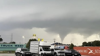 Un tornado desciende de nubes de tormenta oscuras sobre un estacionamiento con vehículos y edificios, incluyendo uno de lavado de camiones y un contenedor de envío
