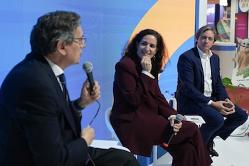 Tres ponentes, dos hombres y una mujer, sentados en un escenario azul y amarillo. Un hombre habla por micrófono. La pantalla muestra el título del evento. Público en primer plano