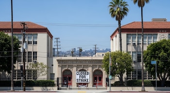 Vista frontal de una escuela pública en Los Ángeles con un cartel "CLOSED FOR STRIKE" en puertas rojas. El edificio es de color crema con techos de tejas rojas y está rodeado de palmeras. No hay personas.