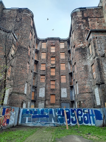 Fachada alta de un edificio antiguo de ladrillos con ventanas tapiadas con madera tras una cerca metálica con pintadas en la parte inferior