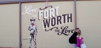 Pared de ladrillo con mural que exhibe las palabras "Love The Fort Worth The Love", una ilustración de vaquero y el hashtag "#LoveTheFort". Dos personas transitan