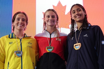 Tres nadadoras sonriendo en un podio. Visten chaquetas deportivas y exhiben una medalla plateada, una dorada y una de bronce