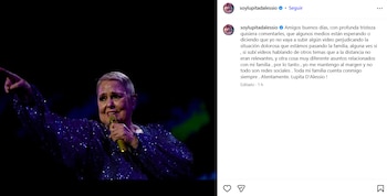 Lupita D'Alessio, con cabello corto y un traje morado brillante, sostiene un micrófono dorado y apunta hacia adelante en un escenario oscuro